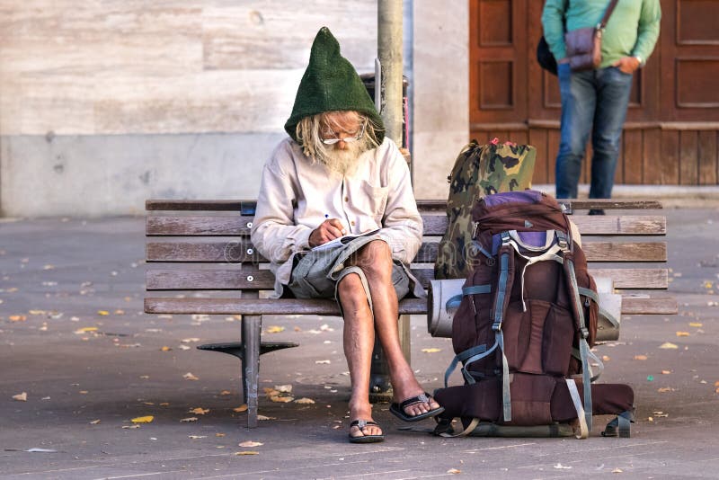 A senior man backpacker sitting on a square bench, is writing down notes on a notepad wearing eyeglasses and a gnome wool hat. Ancona, Italy - September 27th stock photos