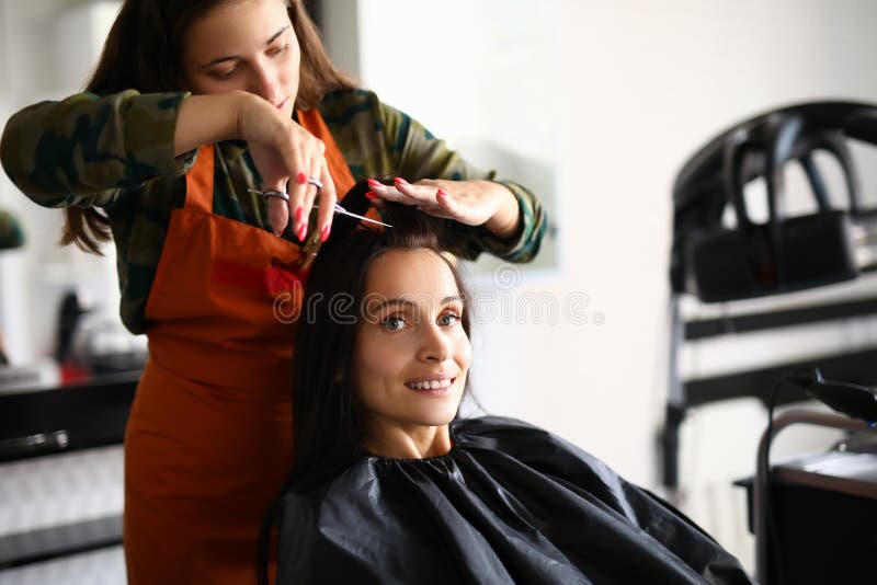Beautiful woman hold strand of hair and cut bang with scissors at workplace. Client sit on chair in cloak and smile. Beautiful women hold strand of hair and cut royalty free stock photos