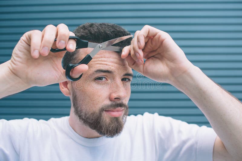 Confident man is standing and holding part of hair in one hand and scissors in another one. He is going to cut off bang. Man is looking at camera. Isolated on royalty free stock photos