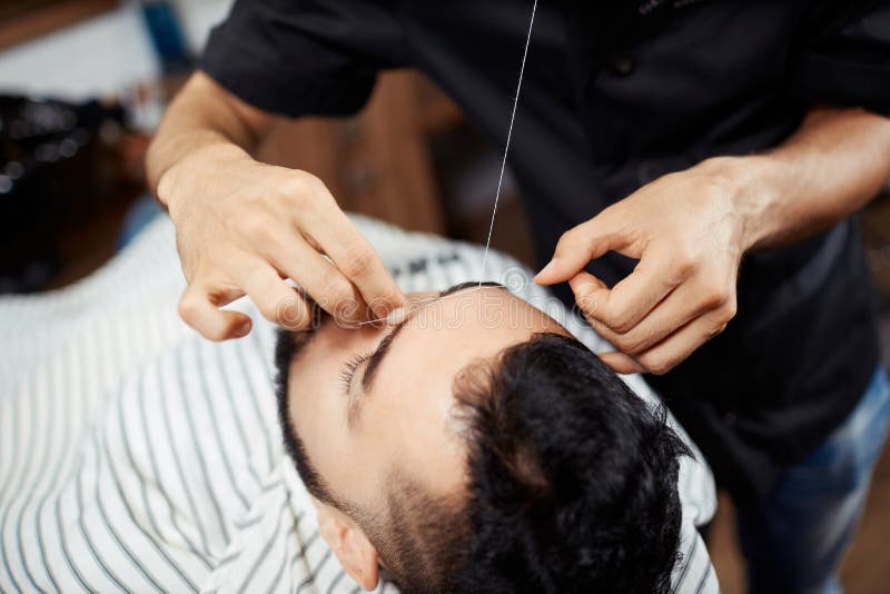 Crop barber threading eyebrows of client. Anonymous man doing eyebrow threading to young man lying in chair with eyes closed stock photo