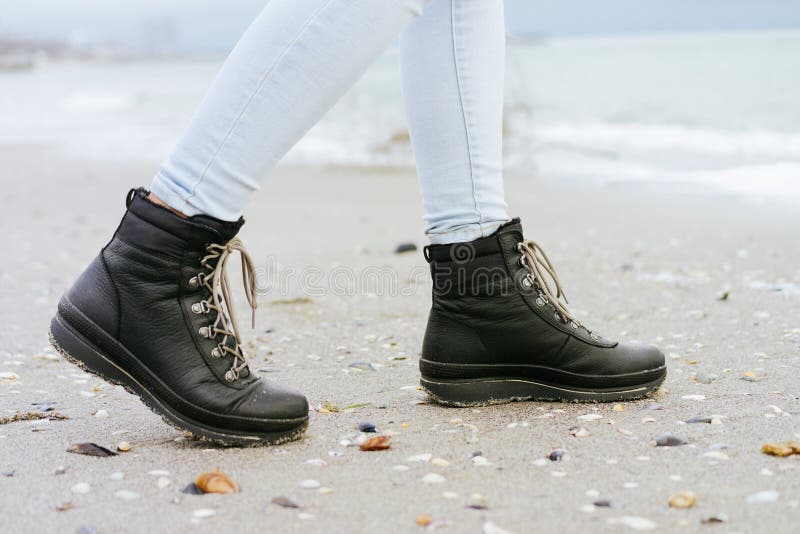 Female feet in blue jeans and black winter boots are on the beackground. Female feet in blue jeans and black winter boots are on the beach sand stock image