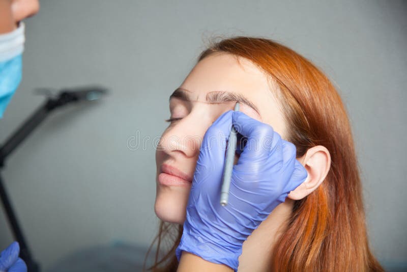 Master on the eyebrows grafted eyebrow. The master on the eyebrows draws the shape of the grafted eyebrow shape in a blue medical disposable gloves red-haired royalty free stock images