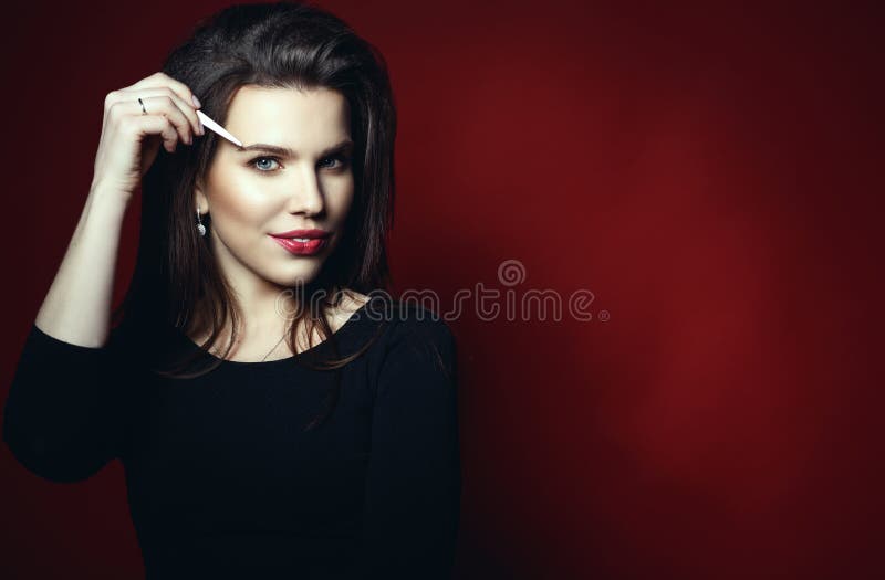 Portrait of beautiful smiling eyebrow master holding tweezers at her eyebrow pretending to pluck it. Isolated on red background. Beauty sphere concept. Studio stock photo