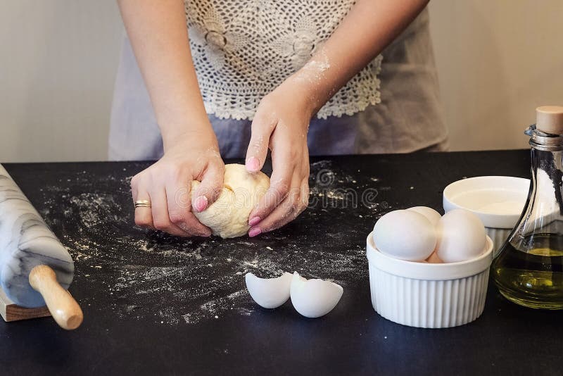 A woman in an apron kneads the dough with her hands on a black wooden table. Ingredients for dough eggs, vegetable oil, flour. On royalty free stock photos