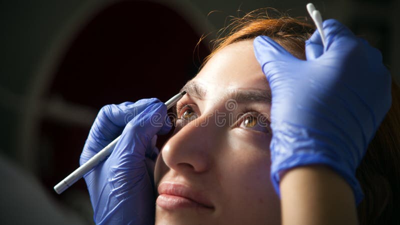 Woman paint her eyebrow. A young red-haired woman paint her eyebrow with a dark master paint to achieve the perfect shape in medical gloves of blue stock photos