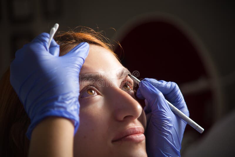 Woman paint her eyebrow. A young red-haired girl dye her eyebrow tassel with a dark master paint to achieve the perfect shape in disposable medical gloves of royalty free stock image