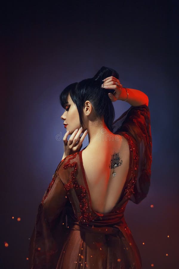 Young girl with dark blue hair and a bang in a long red dress with open naked bare back. a tatoo picture lotus with a. Shiny piercing. hands near the face and royalty free stock image